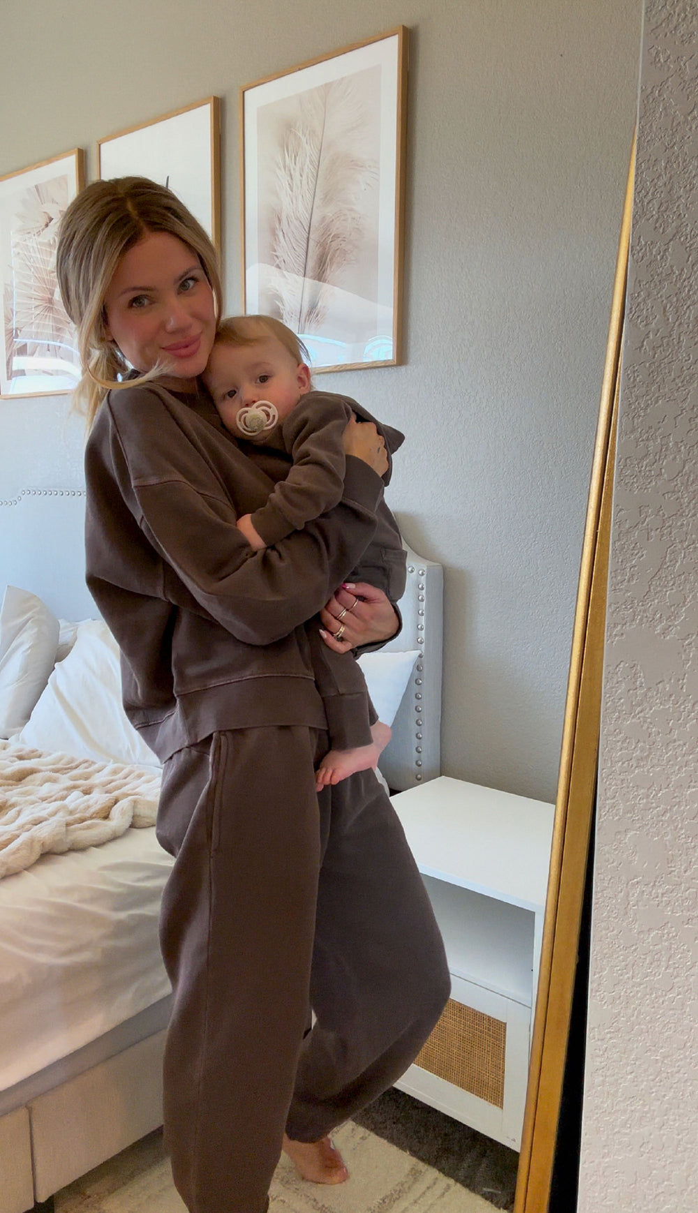 Woman holding a baby in matching brown outfits in a bedroom setting.