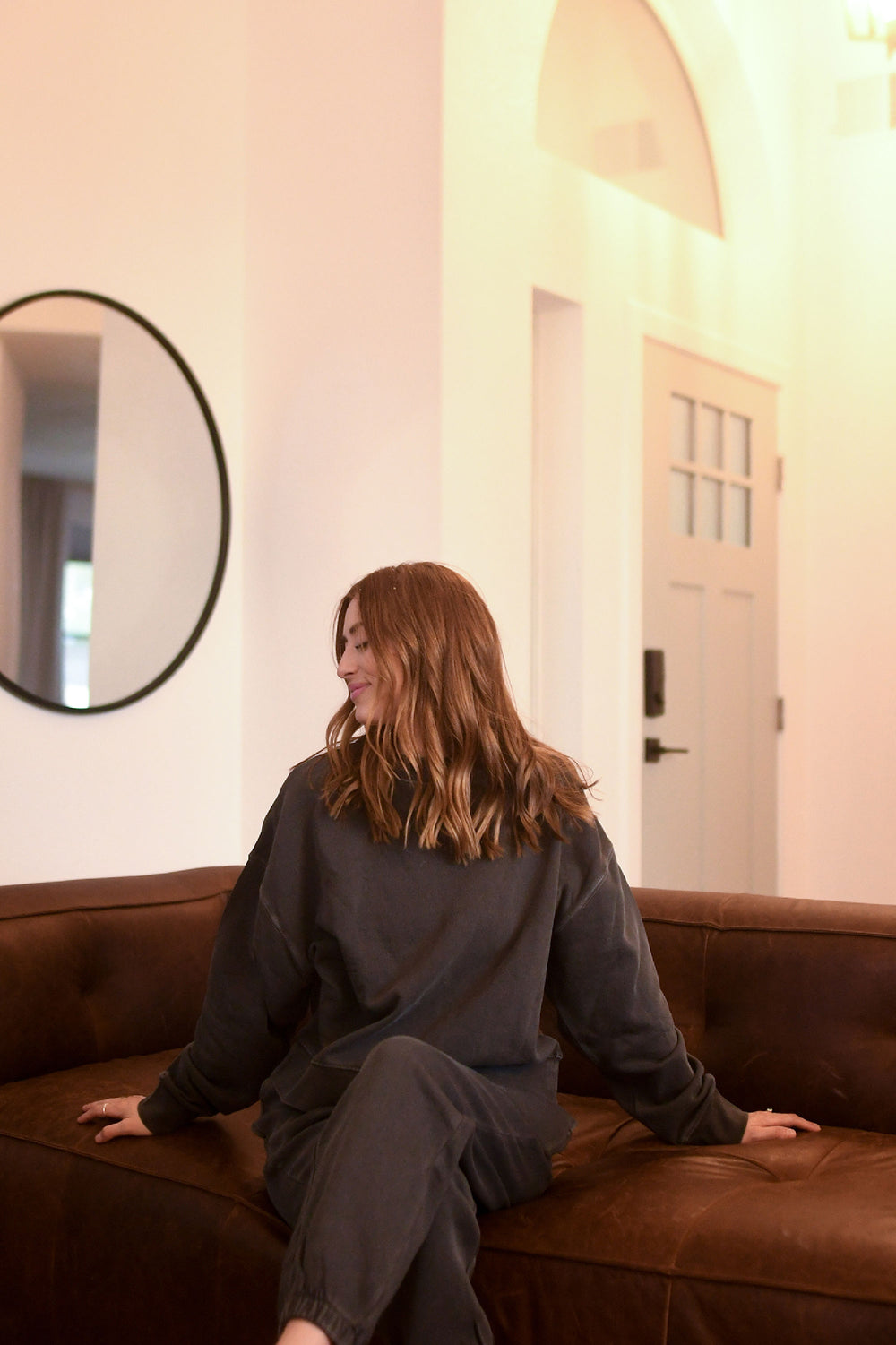 Woman sitting on a brown couch in a room with a mirror and door.