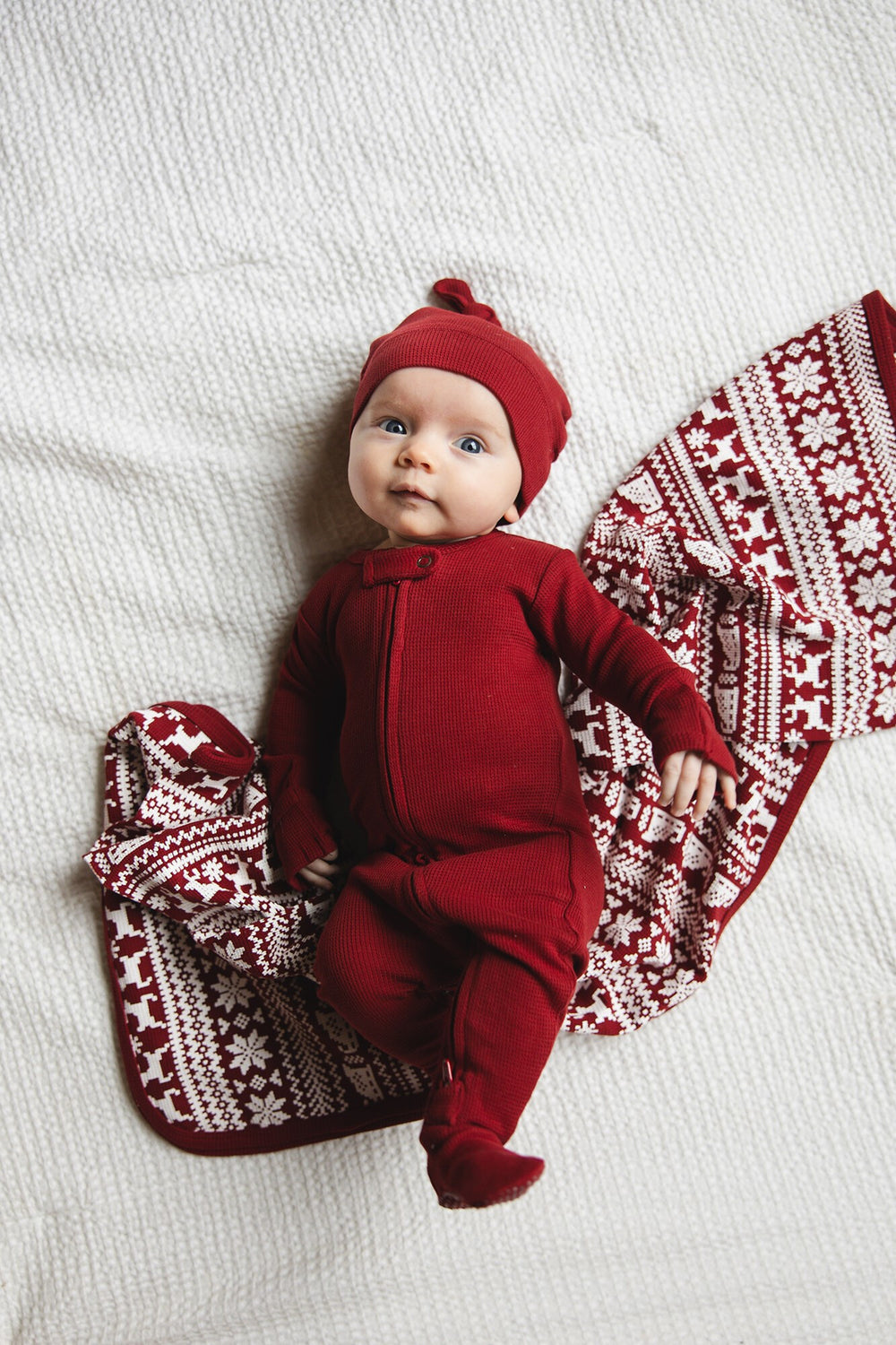 Baby in red outfit with matching hat and patterned blanket on a white background
