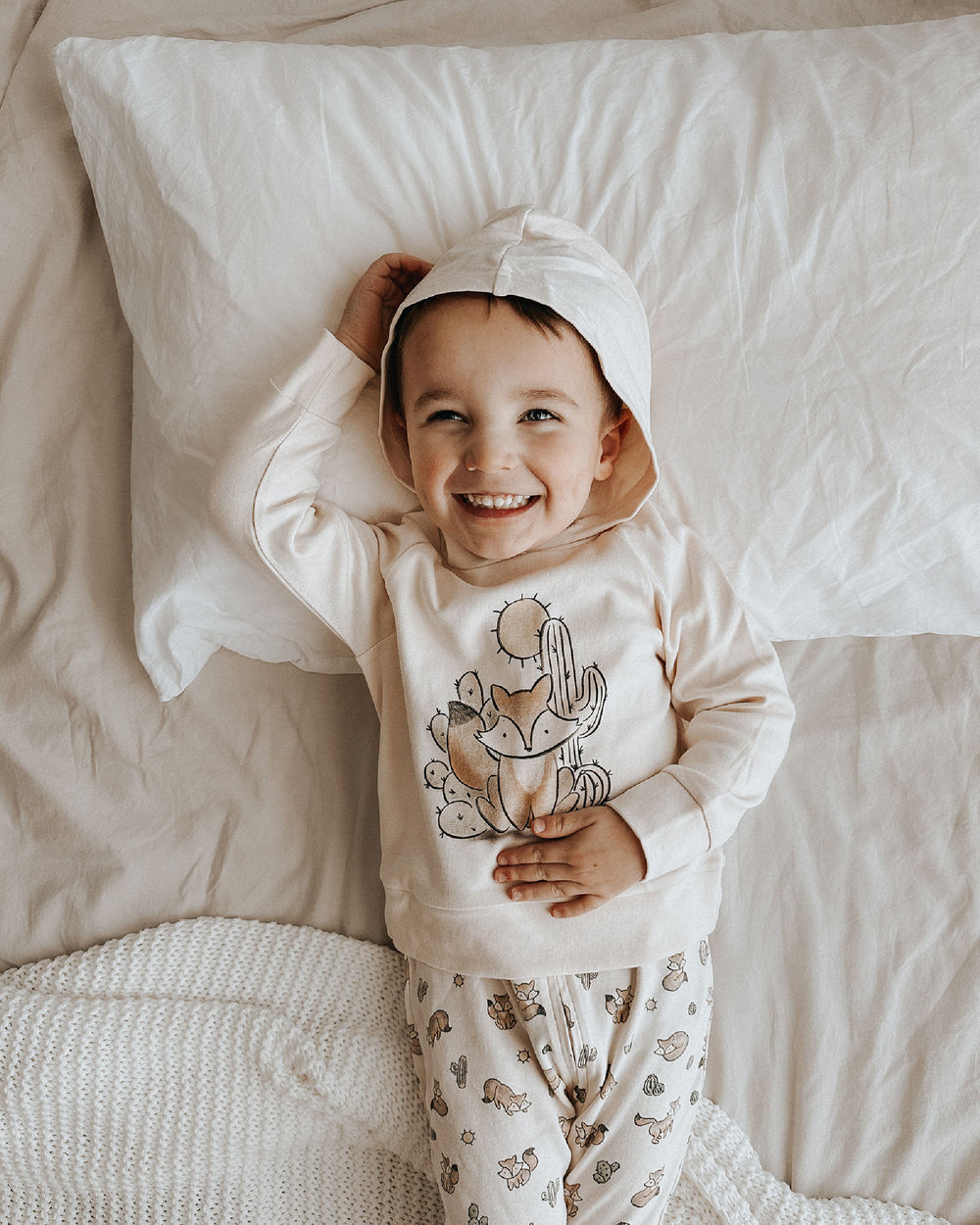 Child wearing a white hoodie with a fox design, lying on a bed.