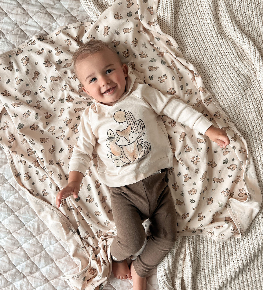 Baby lying on a patterned blanket wearing a white shirt with a design and brown pants.