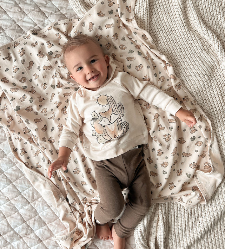 Baby lying on a patterned blanket wearing a white shirt with a design and brown pants.