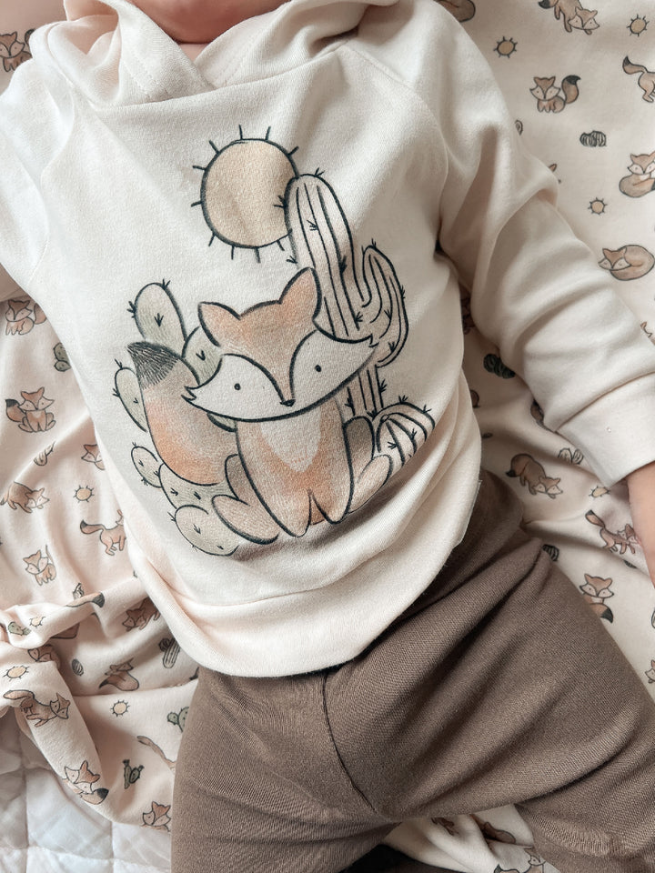 Child wearing a white hoodie with a fox and cactus design on a patterned surface