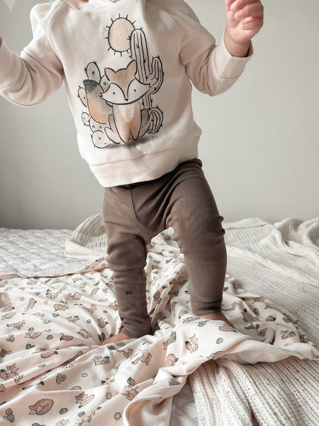 Child wearing a sweatshirt with a fox and cactus design on a bed.