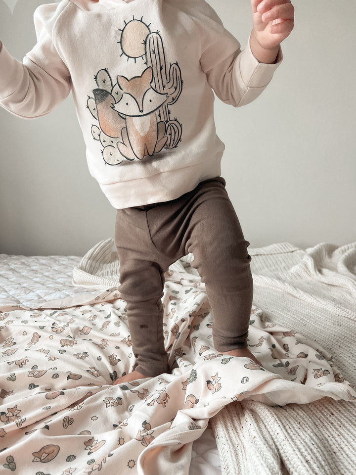 Child wearing a sweatshirt with a fox and cactus design on a bed.