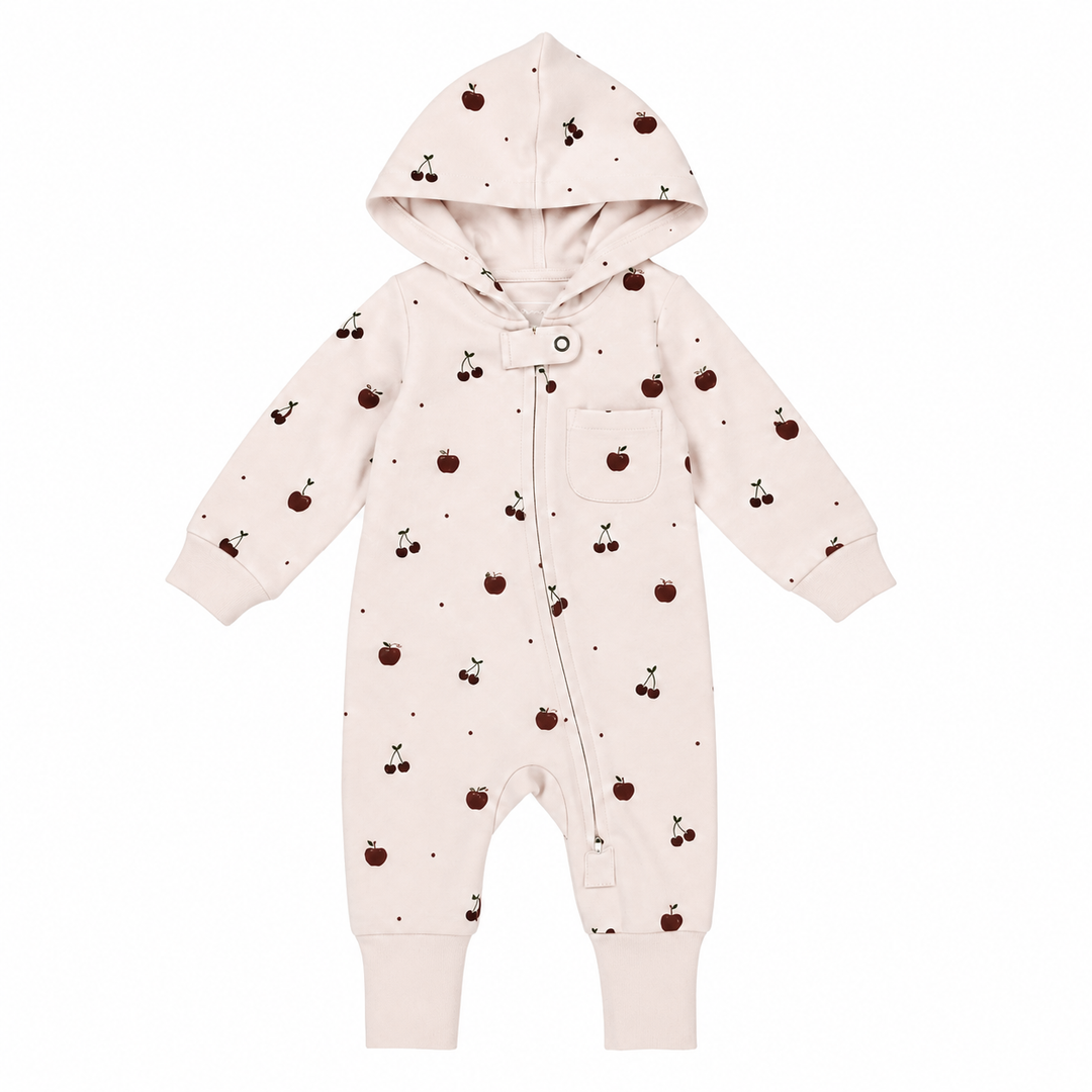 A baby hooded one-piece romper in a very pale blush pink, featuring a small all-over print of dark red cherries and tiny black dots, with a snap-button front and a chest pocket.