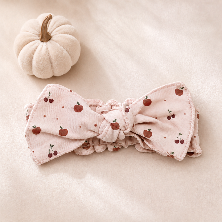A light pink fabric headband with a knotted bow, decorated with a pattern of red cherries and small red dots, shown on a plain white background.
