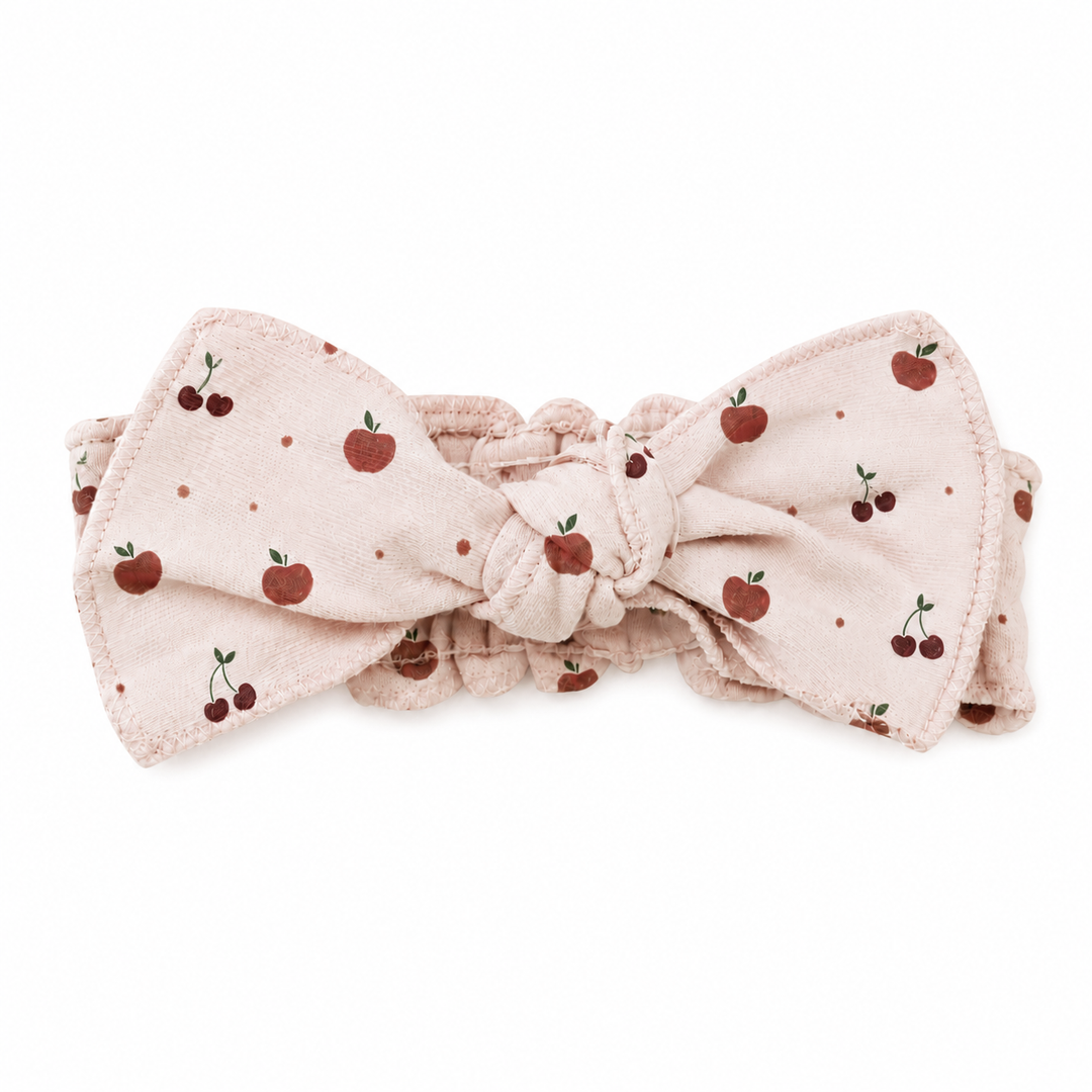 A light pink fabric headband with a knotted bow, decorated with a pattern of red cherries and small red dots, shown on a plain white background.