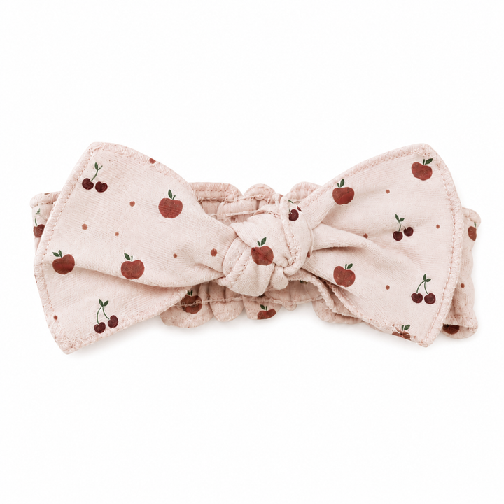 A light pink fabric headband with a knotted bow, decorated with a pattern of red cherries and small red dots, shown on a plain white background.