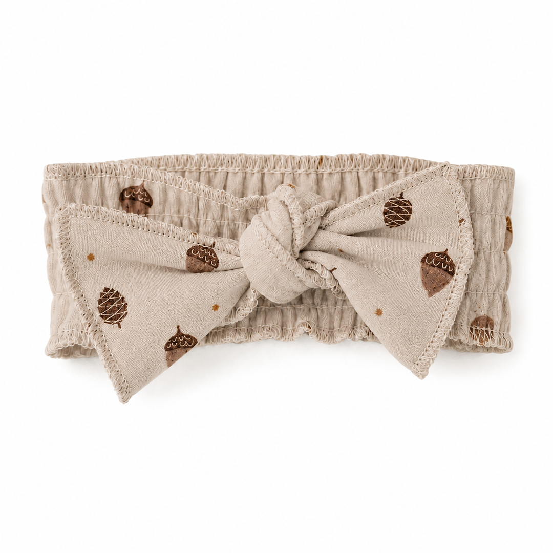 A beige fabric headband with a large front bow, featuring a brown acorn and pinecone pattern, photographed on a solid white background.