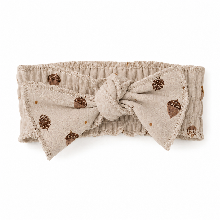 A beige fabric headband with a large front bow, featuring a brown acorn and pinecone pattern, photographed on a solid white background.