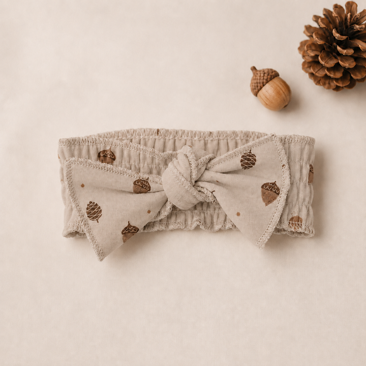 A beige fabric headband with a large front bow, featuring a brown acorn and pinecone pattern.