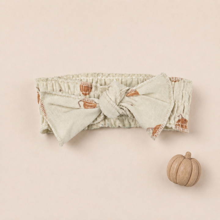 A cream-colored fabric headband with a large bow, featuring a pattern of orange pumpkins.