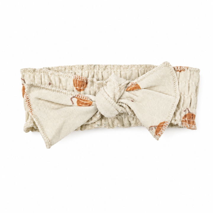 A cream-colored fabric headband with a large bow, featuring a pattern of orange pumpkins and small brown animals, shown on a plain white background.