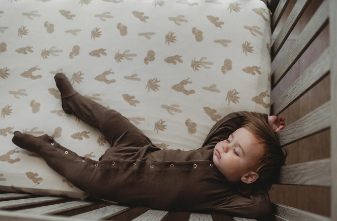 Child sleeping in crib, wearing solid brown organic footie. Mattress in the crib is fitted with our crib sheet in the buttercream cactus color option.