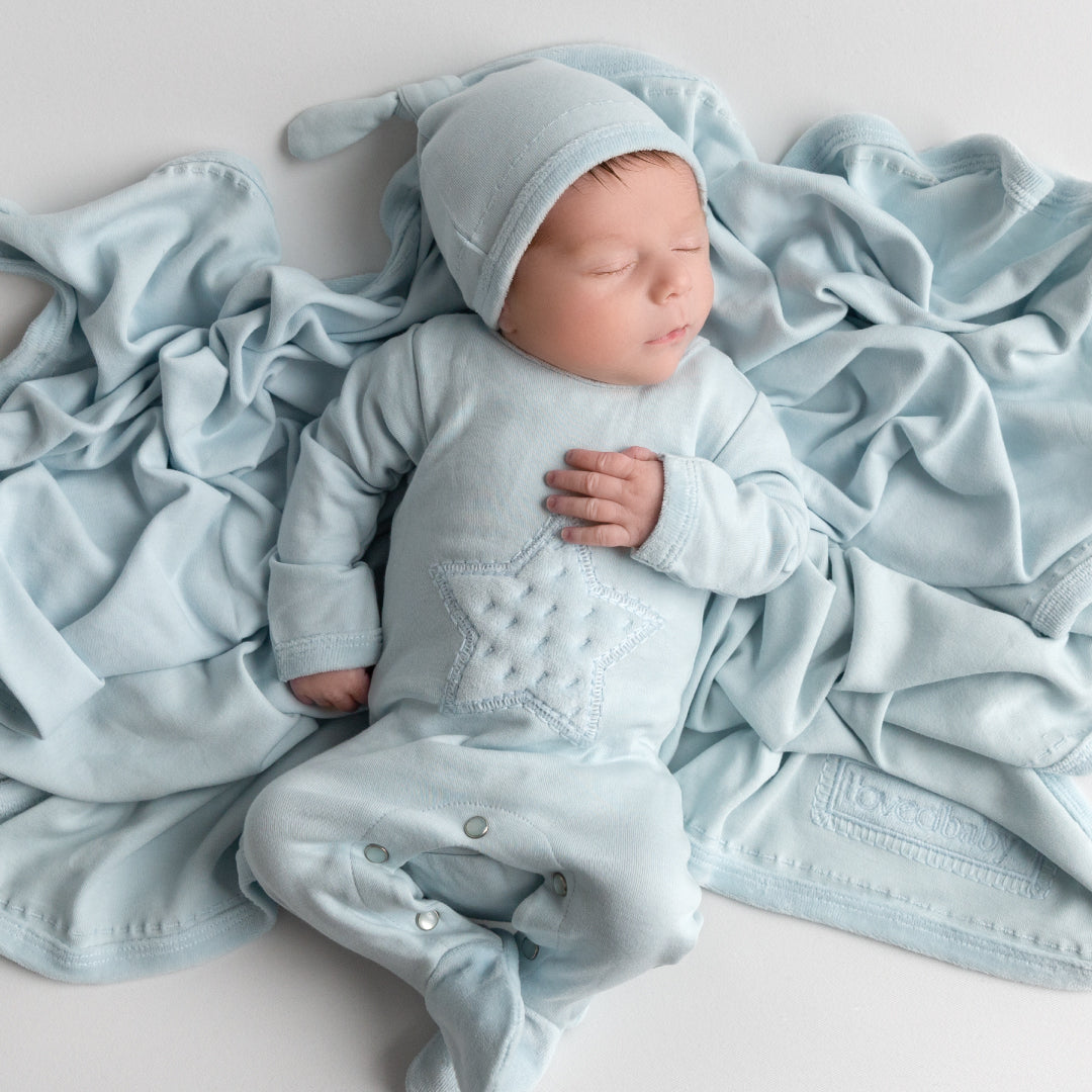 Image of boy sleeping, laying on moonbeam colored blanket from Velveteen collection. Wearing star appliqué footie from same collection and matching knotted hat.