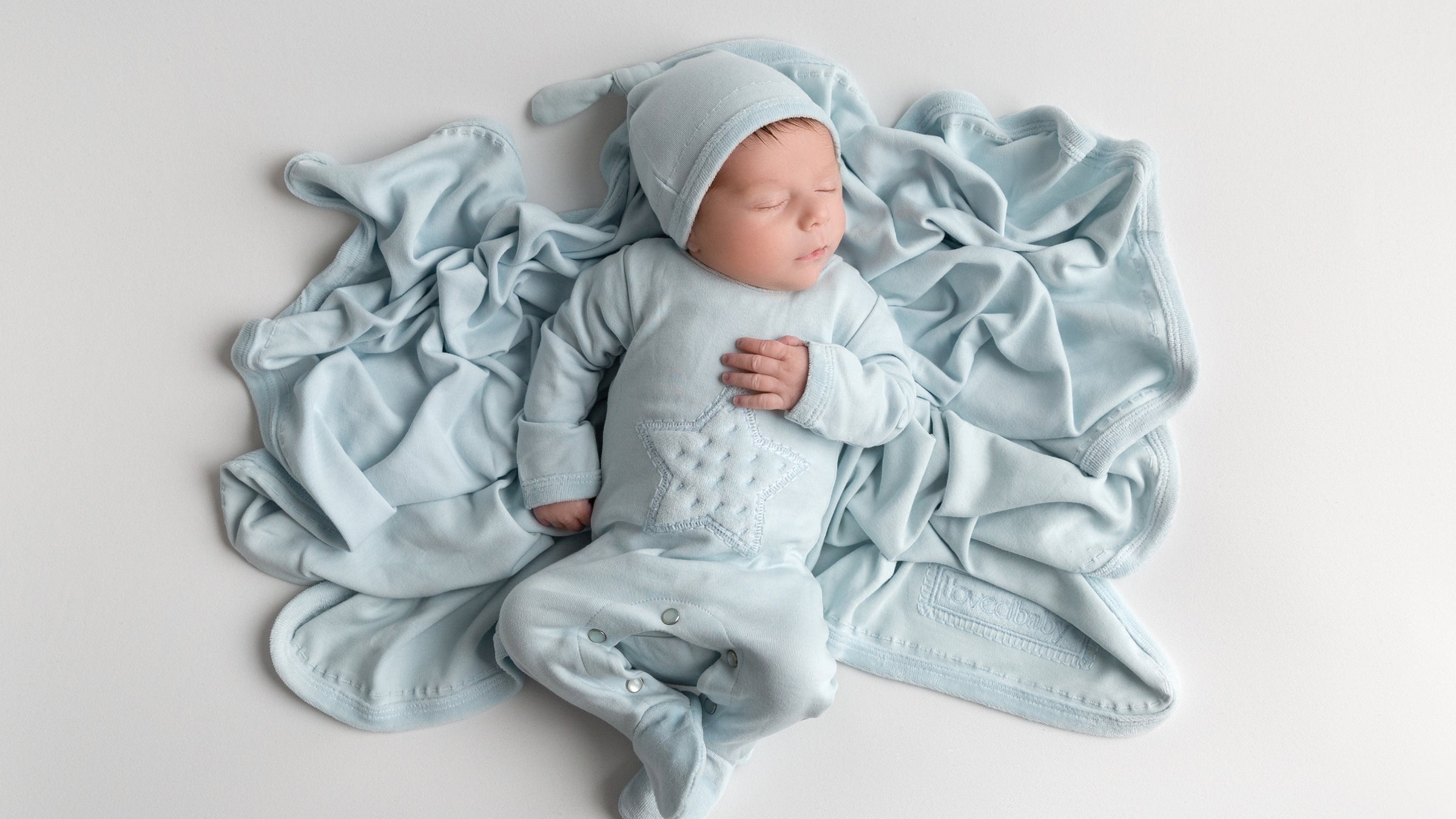 Image of boy sleeping, laying on moonbeam colored blanket from Velveteen collection. Wearing star appliqué footie from same collection and matching knotted hat.