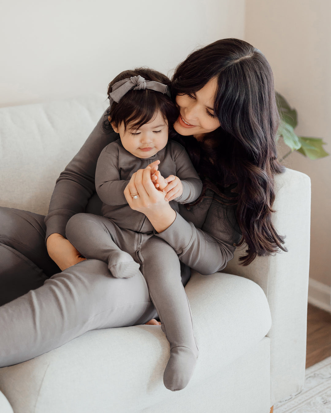 Woman sitting on couch wearing women's lounge set in gray. baby sitting on lap wearing gray zipper footie and smocked headband