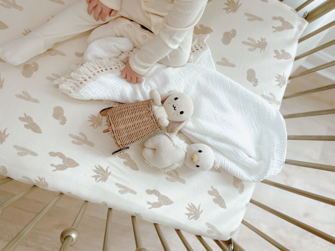 Child sitting in crib, wearing solid white organic footie. Mattress in the crib is fitted with our crib sheet in the buttercream cactus color option.