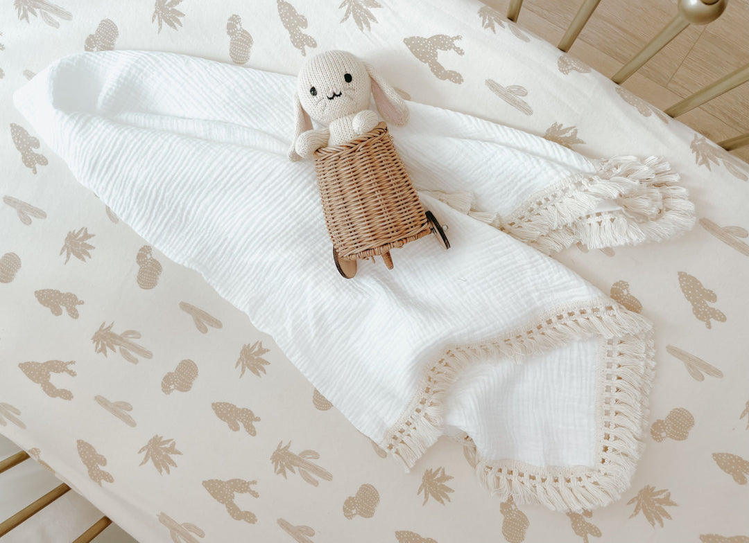A doll laying on top of a white blanket in a crib. Mattress in the crib is fitted with our crib sheet in the buttercream cactus color option.