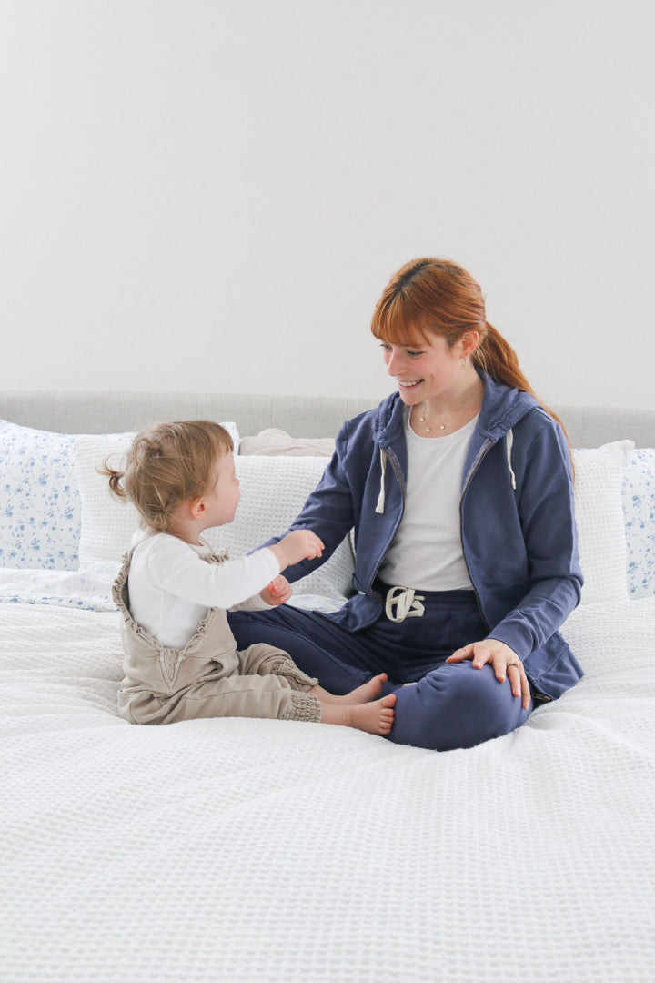 Woman wearing Indigo colored jacket and jogger, interacting with a toddler wearing Oatmeal romper.
